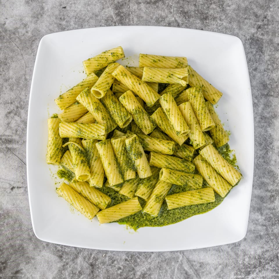 A white square plate filled with rigatoni pasta coated in vibrant green pesto sauce.