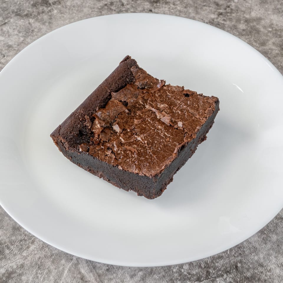 A rich, dark chocolate brownie with a crinkled top sits on a white plate.