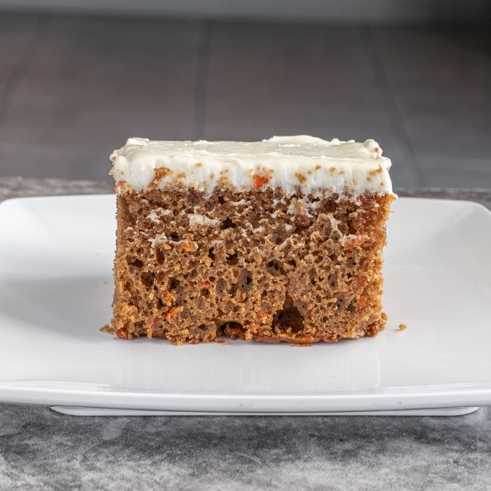 A square slice of carrot cake with white frosting sits on a white rectangular plate.