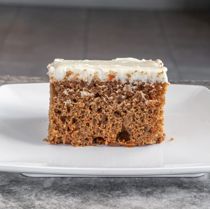 A square slice of carrot cake with white frosting sits on a white rectangular plate.