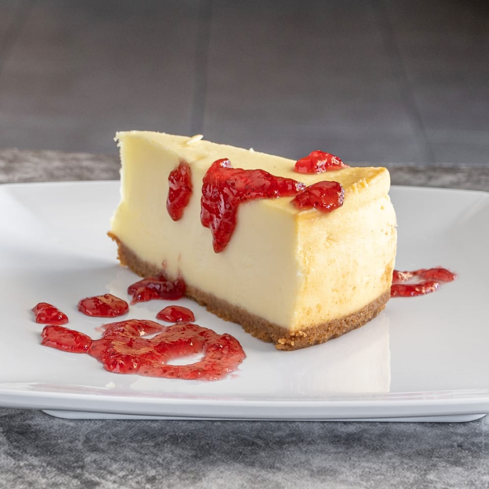 A slice of Ameci cheesecake with a graham cracker crust, topped with bright red strawberry sauce.